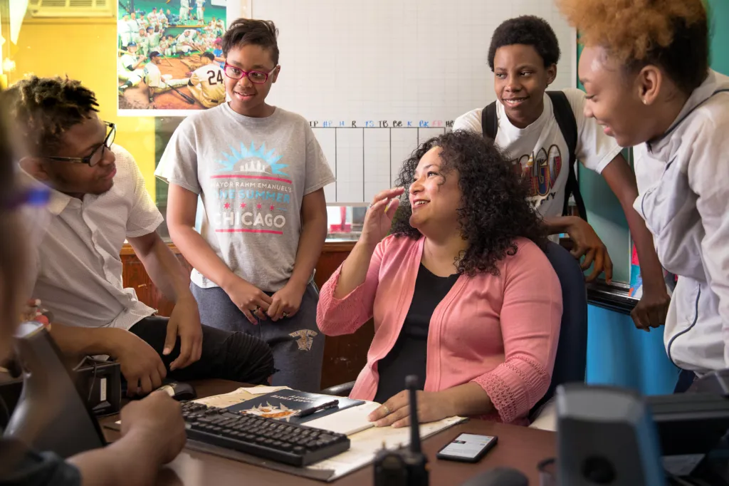 group of happy students gathering around teacher at desk