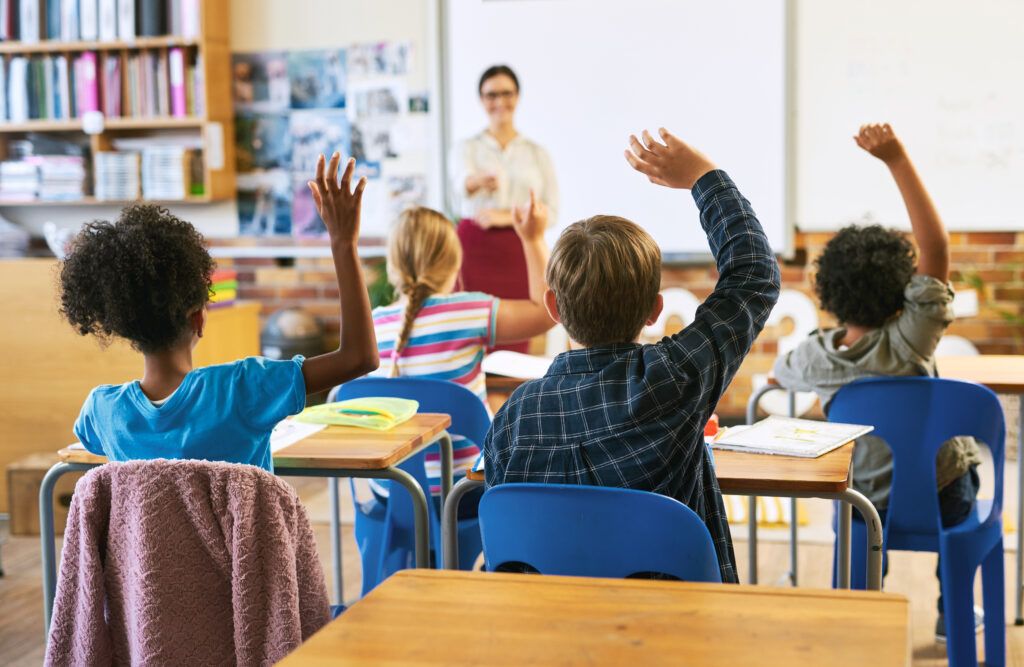 group of children sitting at desks in a classroom with hands raised