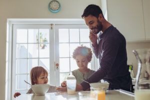 Man standing at the breakfast table talking over mobile phone with kids eating breakfast.
