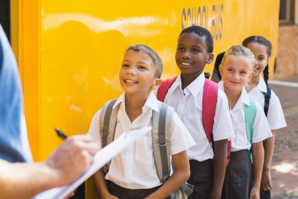 students lining up outside school bus to be checked off by school staff member with clipboard 