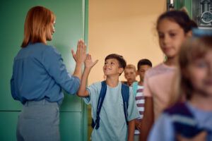 Happy boy giving high-five to his teacher while getting int he classroom with his classmates.