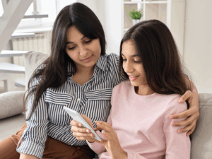 mother and teenage daughter sitting next to each other on couch looking at mobile phone