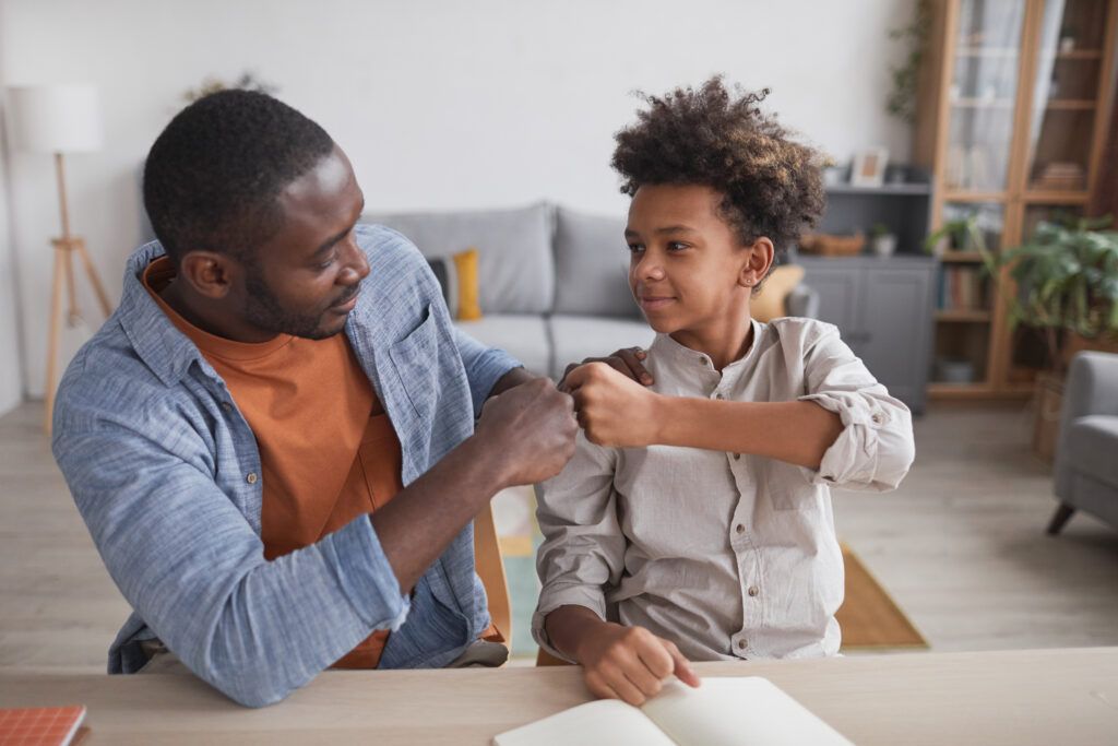 Portrait of father fist bumping smiling son while doing homework together at home