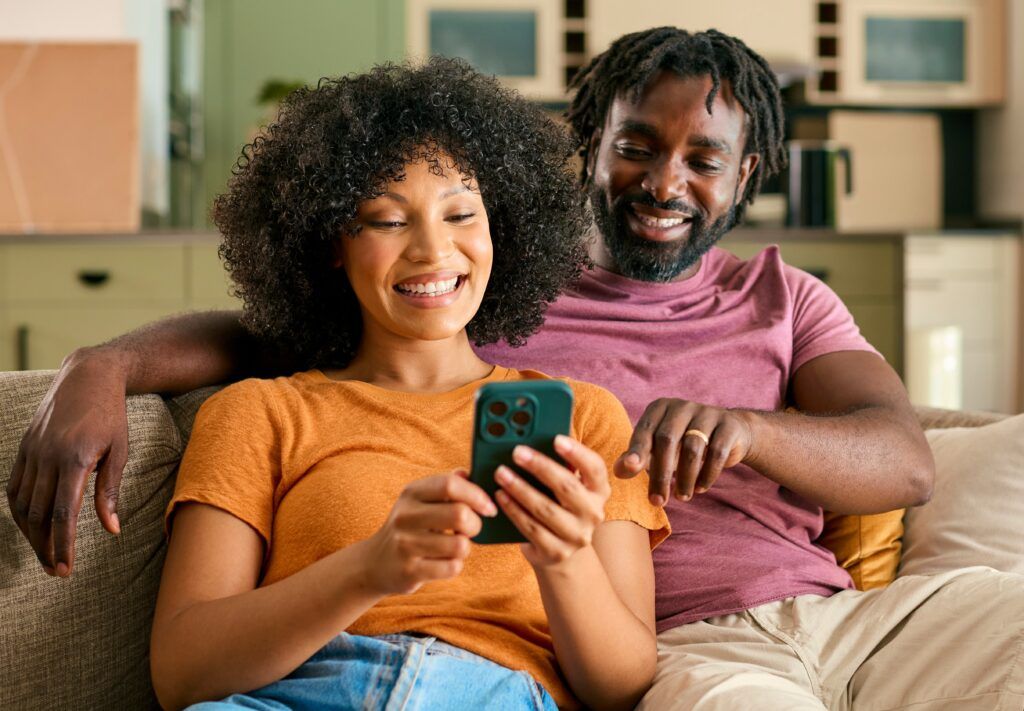 Couple Sitting On Sofa At Home Looking At Mobile Phone Together