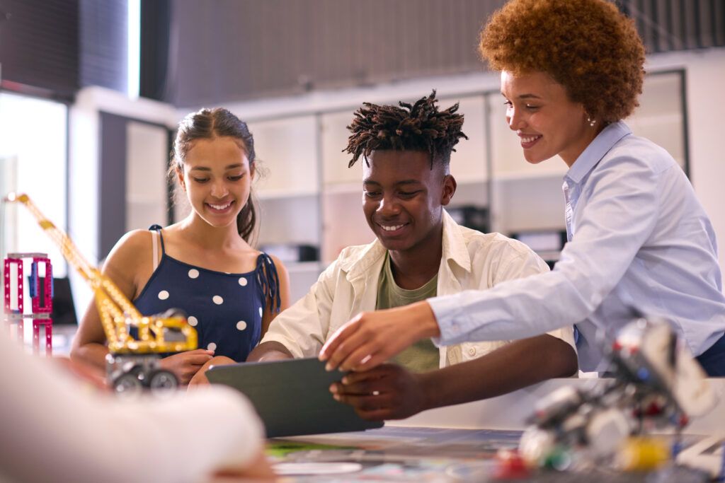 Female High School Teacher With Digital Tablet Helping Students In STEM Technology Lesson