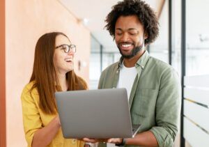 Two smiling colleagues talking over laptop in hallway