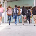 Group of school children wearing backpacks running down a covered outdoor school hallway toward a building entrance, captured from behind during the school day.