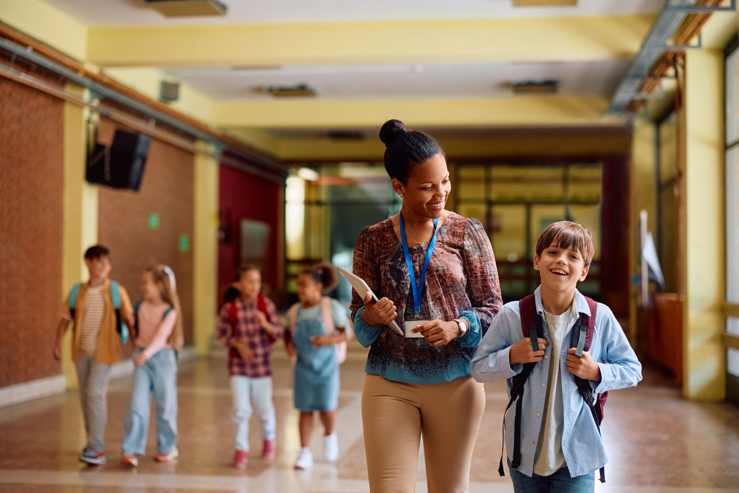 A smiling teacher walks through a school hallway with a group of young students wearing backpacks.