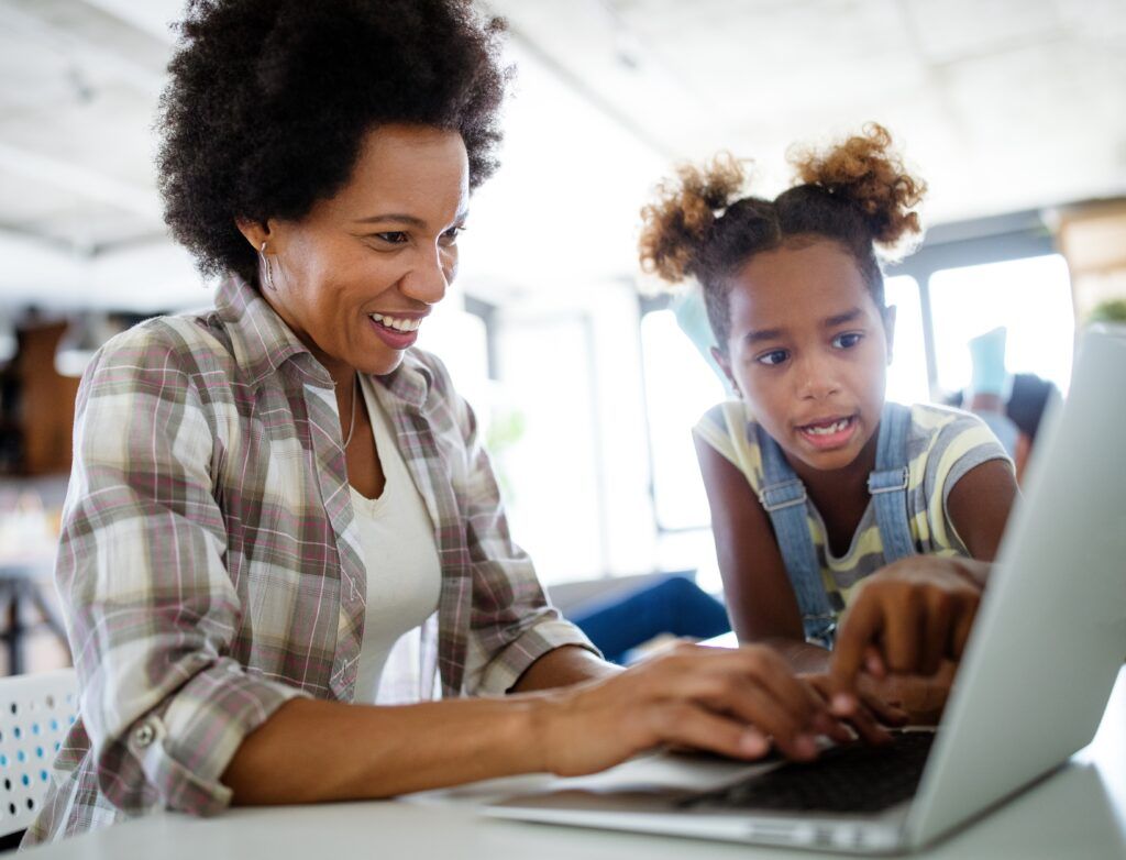Smiling mother and young daughter using a laptop together at home, child pointing at screen while learning online, family bonding over technology, casual indoor setting with natural light, education and digital learning concep