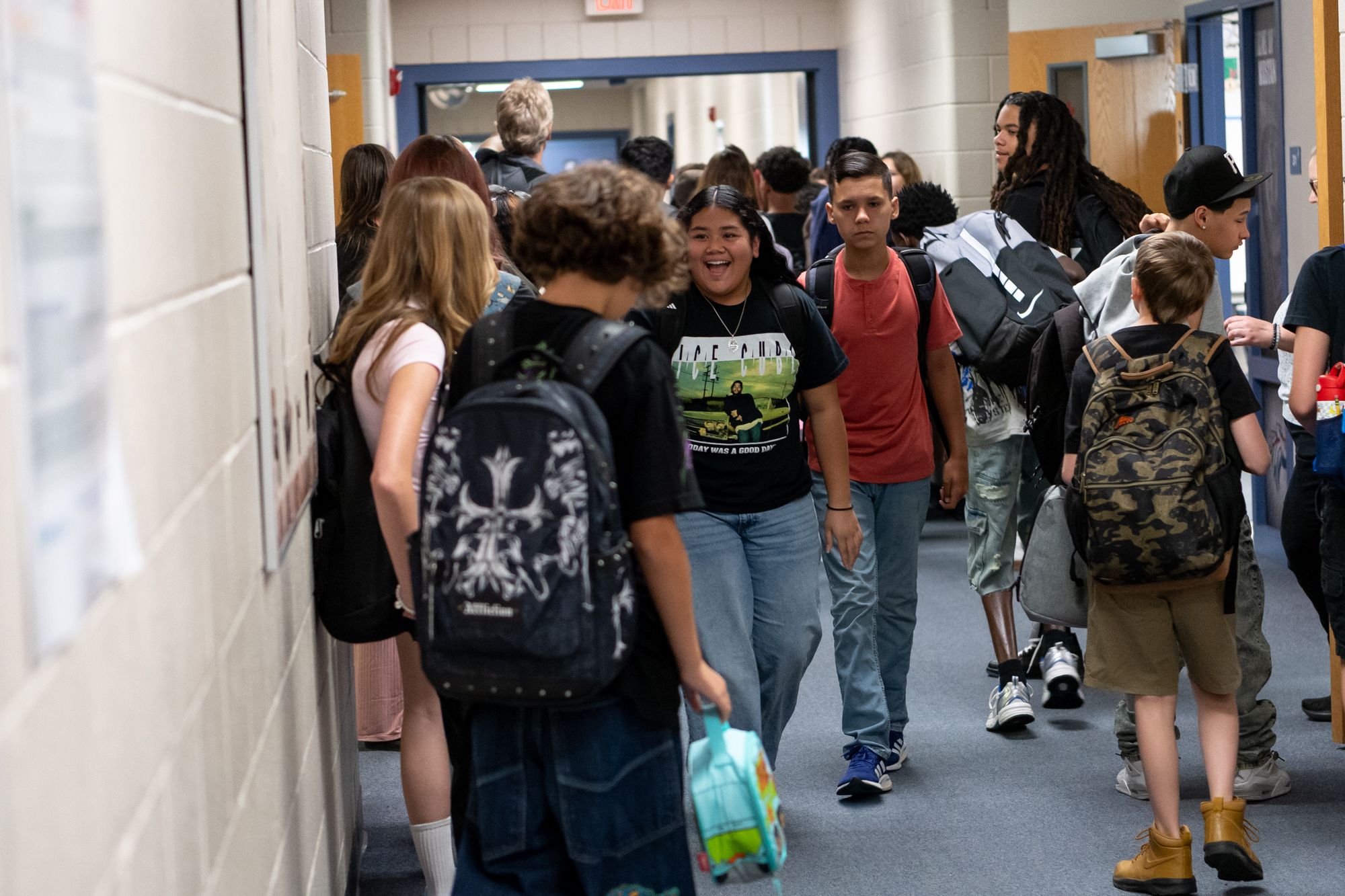 Students walk through a busy school hallway between classes, carrying backpacks and connecting with classmates.