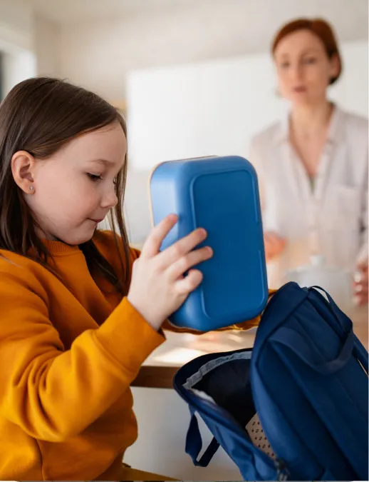 Young girl placing a blue lunch container into a backpack while an adult looks on in a home kitchen or dining area.