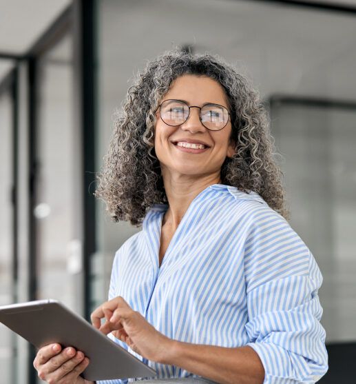Smiling woman with curly gray hair and glasses holding a tablet in a modern office setting.