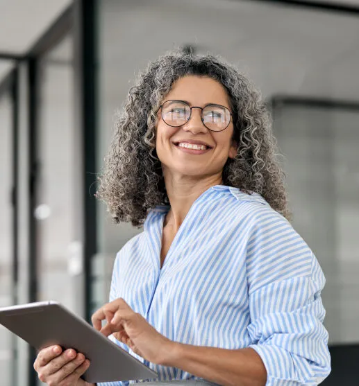 Smiling woman with curly gray hair and glasses holding a tablet in a modern office setting.