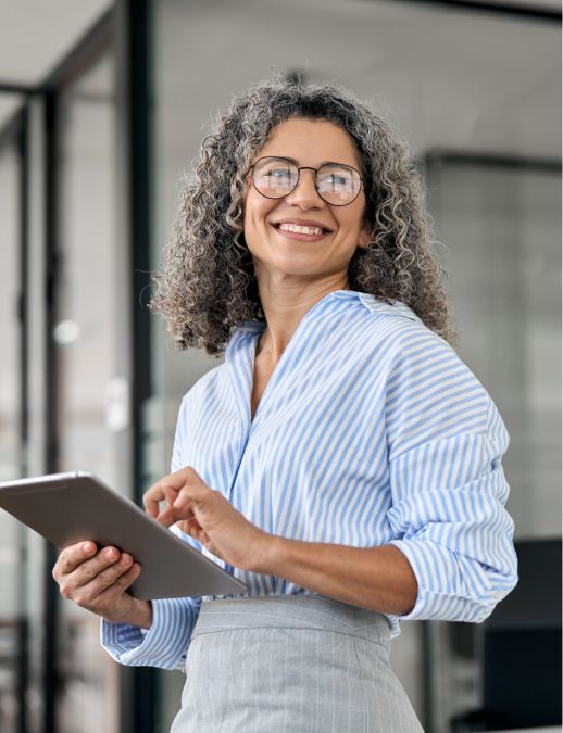 Smiling woman with curly gray hair and glasses holding a tablet in a modern office setting.