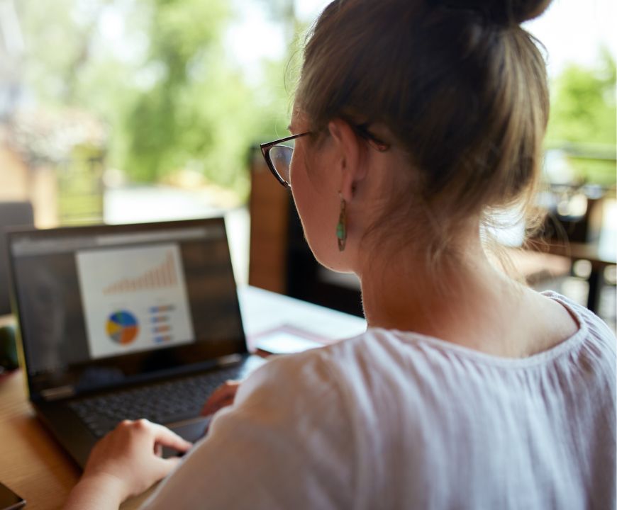 Woman wearing glasses looks at a laptop screen displaying charts and graphs while working at a desk in a bright indoor setting.