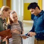 School staff member showing a tablet to a woman and teenage girl while talking together in a bright school hallway.