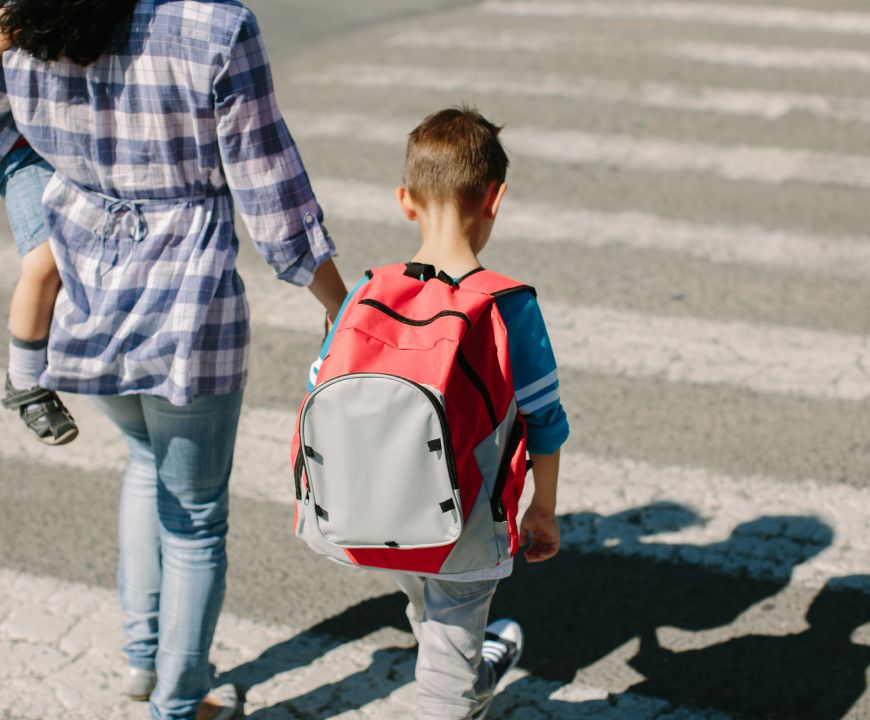 Adult holding a young child’s hand while walking with a school-age boy wearing a red backpack across a crosswalk outdoors.
