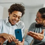 Teen boy and parent smiling while looking at a smartphone together during a casual conversation in a home setting.