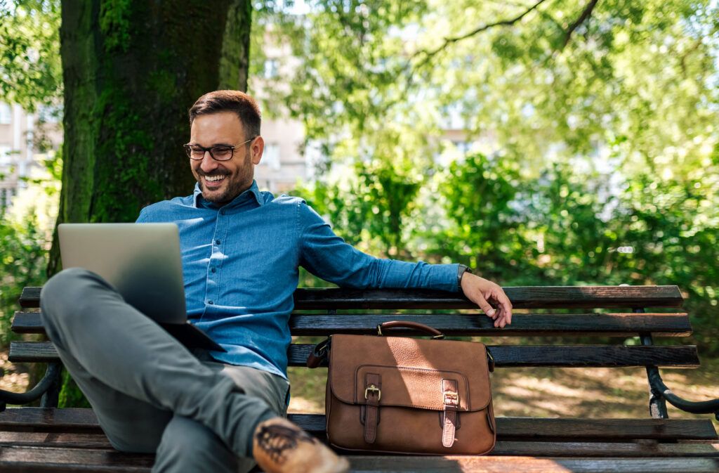 Casual man, sitting cross-legged on the bench, resting his handbag next to him.