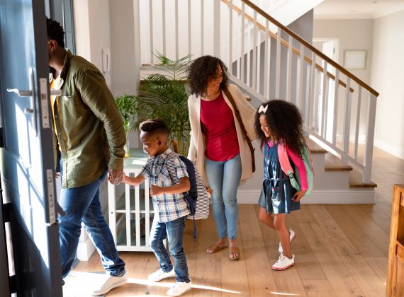 Family with two children wearing backpacks walking out door of home