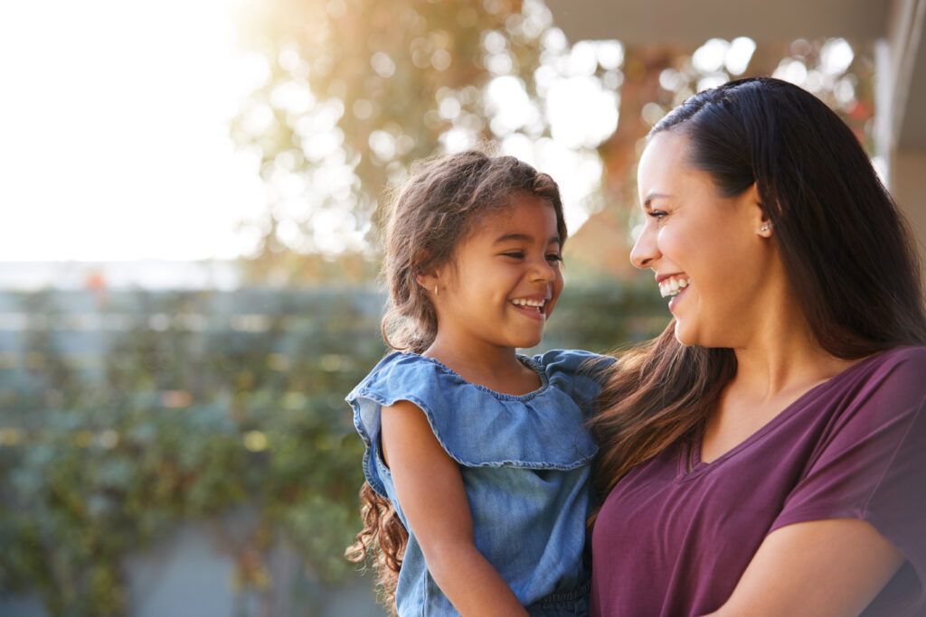A smiling parent holds a young child outdoors as they look at each other and laugh.