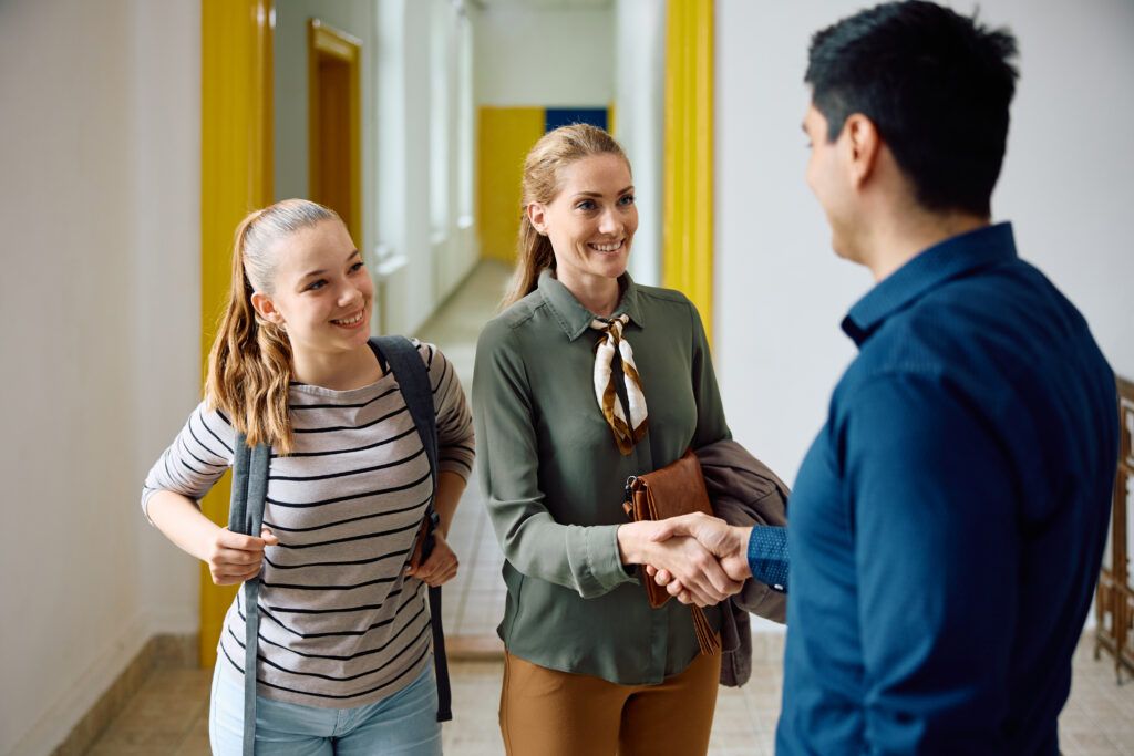 Happy mother of teenage girl shaking hands with high school principal in hallway