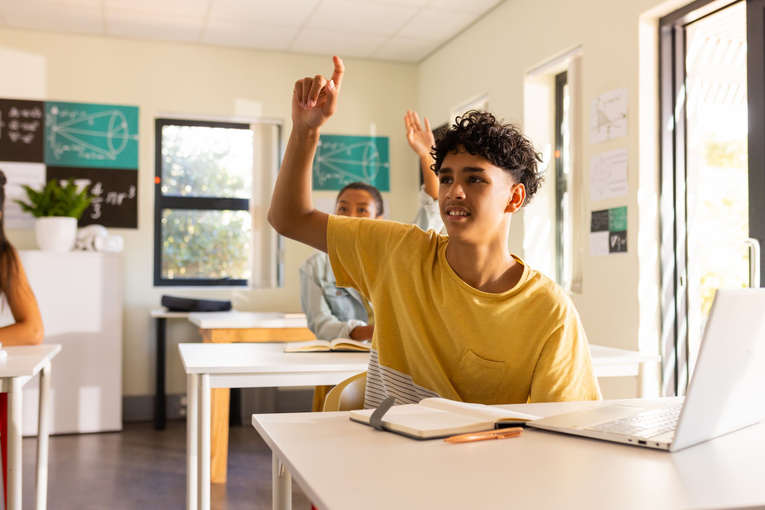 teenage boy raising hand while sitting at desk with laptop